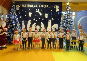 Dzieci śpiewają piosenkę, w tle dekoracja świąteczna.